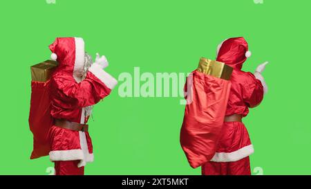 Saint Nick mit vollem Sack per Anhalter vor der Kamera, um die Welt am weihnachtsabend zu bereisen. Junger Mann, der den weihnachtsmann mit Geschenken und Spielzeug in der Tasche darstellt, Greenscreen-Copyspace. Stockfoto