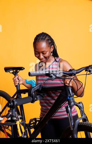 Vor gelbem Hintergrund montiert die aktive gesunde Frau ihr Fahrrad auf einem Reparaturständer zur Befestigung. Bild einer sportlichen schwarzen Frau, die die Höhe ihres Fahrrads ändert, um die regelmäßige Wartung vorzubereiten. Stockfoto