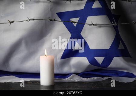 Brennende Kerze mit Stacheldraht und großer Flagge Israels auf dunklem Hintergrund. Holocaust-Gedenktag Stockfoto
