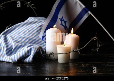 Gefangenenuniform, brennende Kerzen, Stacheldraht und Flagge Israels auf dunklem Hintergrund. Holocaust-Gedenktag Stockfoto