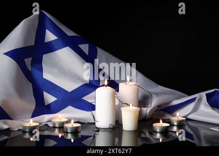 Brennende Kerzen mit Stacheldraht und großer Flagge Israels auf dunklem Hintergrund. Holocaust-Gedenktag Stockfoto
