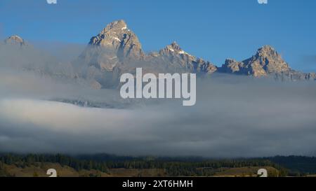 Ein atemberaubender Blick auf die Grand Teton Bergkette mit tief liegenden Wolken und einem klaren blauen Himmel in Teton County, Wyoming, USA Stockfoto