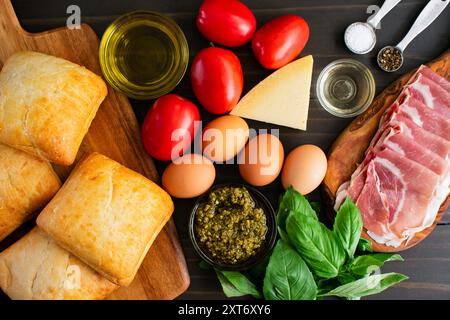 Italienische Sandwichzutaten auf Holztisch: Pflaumentomaten, Eier, Pesto, Ciabatta-Brötchen, Parmesankäse, Prosciutto und Basilikum auf dunklem Hintergrund Stockfoto