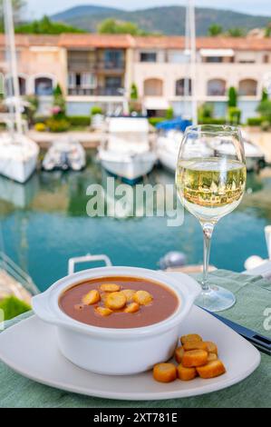 Hausgemachte leckere Fisch- oder Biskuitsuppe mit Croutons, serviert im Freien mit einem Glas kalten Weißwein und Blick auf Yachtboote und Häuser von Port Grimaud, Fren Stockfoto