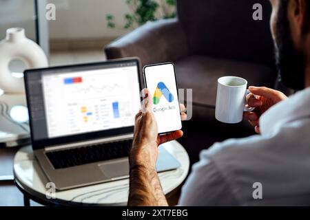 Mann, der ein Mobiltelefon mit Google Ads auf dem Bildschirm und im Hintergrund einen Computer mit Anzeigenstatistiken verwendet. Rosario, Argentinien - 23. Juli 2024. Stockfoto