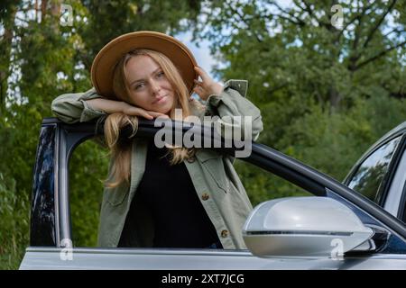 Blonde Frau mit Hut, die neben der Autotür wohnt. Junge Touristen erkunden lokale Reisen und machen echte Momente. Wahre Emotionen Ausdruck des Ausgehens und der Erfrischung Entspannen Sie sich an der frischen Luft Stockfoto