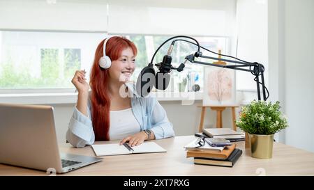 Eine schöne asiatische Podcaster mit roten Locken arbeitet in ihrem Studio, trägt Kopfhörer und spricht ins Mikrofon, sendet oder kündigt an Stockfoto