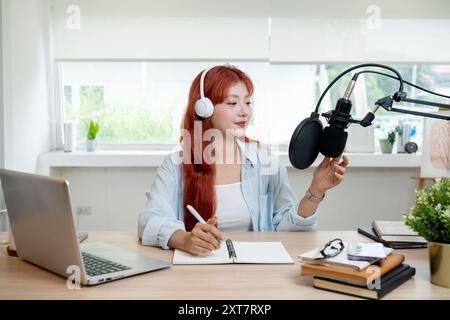 Eine schöne asiatische Podcaster mit roten Locken arbeitet in ihrem Studio, trägt Kopfhörer und spricht ins Mikrofon, sendet oder kündigt an Stockfoto