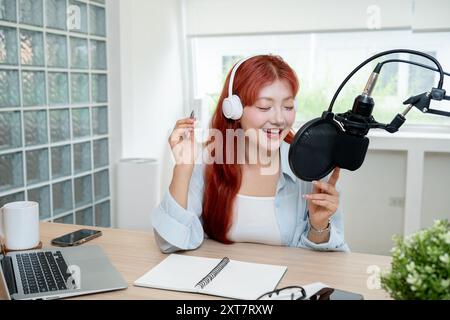 Eine schöne, fröhliche asiatische Podcaster mit rotem lockigem Haar arbeitet in ihrem Studio, trägt Kopfhörer und spricht ins Mikrofon und sendet Stockfoto