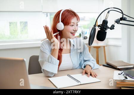 Eine schöne, fröhliche asiatische Podcaster mit rotem lockigem Haar arbeitet in ihrem Studio, trägt Kopfhörer und spricht ins Mikrofon und sendet Stockfoto