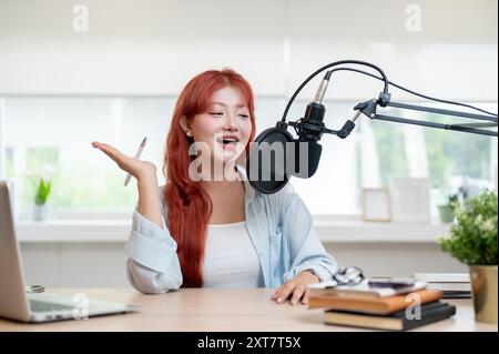 Eine charmante und fröhliche asiatische Podcaster mit rotem lockigem Haar spricht in ein Mikrofon und genießt die Aufnahme ihres Podcasts im Stockfoto