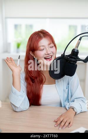 Eine charmante und fröhliche asiatische Podcaster mit rotem lockigem Haar spricht in ein Mikrofon und genießt die Aufnahme ihres Podcasts im Stockfoto