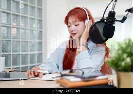 Eine wunderschöne asiatische Podcaster mit roten Locken trägt Kopfhörer und konzentriert sich auf das Lesen des Drehbuchs während der Aufnahme ihres Podcasts. Stockfoto