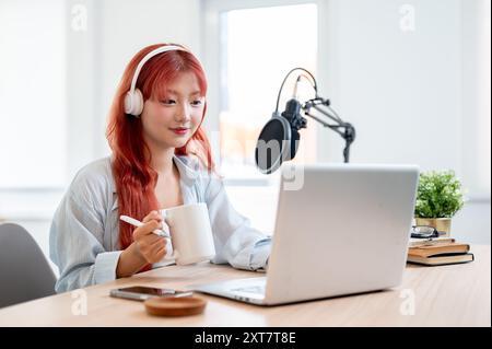 Eine selbstbewusste und schöne asiatische Podcasterin arbeitet in ihrem Heimstudio und genießt einen Kaffee, während sie eine neue Episode ihres Podcasts aufnimmt. Stockfoto