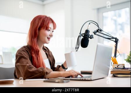 Eine selbstbewusste und schöne asiatische Podcasterin arbeitet in ihrem Heimstudio und genießt einen Kaffee, während sie eine neue Episode ihres Podcasts aufnimmt. Stockfoto