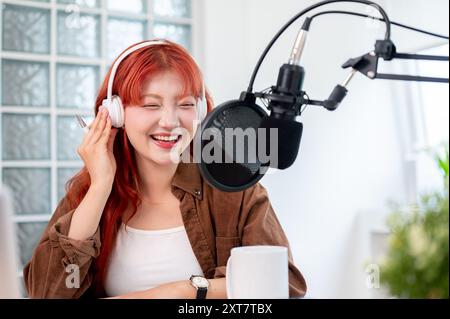 Eine schöne und fröhliche asiatische Podcaster mit rotem lockigem Haar trägt Kopfhörer und genießt es, ihren Podcast im Studio zu leiten. Stockfoto