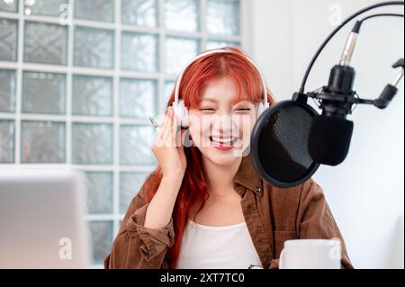 Eine schöne und fröhliche asiatische Podcaster mit rotem lockigem Haar trägt Kopfhörer und genießt es, ihren Podcast im Studio zu leiten. Stockfoto