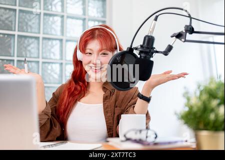 Eine schöne und fröhliche asiatische Podcaster mit roten lockigen Haaren spricht mit geöffneter Hand, trägt Kopfhörer und genießt es, ihre Pod zu leiten Stockfoto