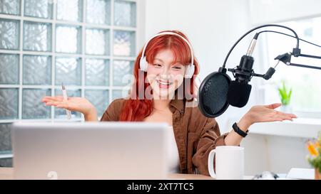 Eine schöne und fröhliche asiatische Podcaster mit roten lockigen Haaren spricht mit geöffneter Hand, trägt Kopfhörer und genießt es, ihre Pod zu leiten Stockfoto