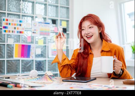 Eine fröhliche asiatische Grafikerin mit roten Locken lächelt in die Kamera, hält eine Kaffeetasse und sitzt an ihrem Schreibtisch im Büro, sur Stockfoto