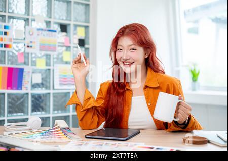 Eine fröhliche asiatische Grafikerin mit roten Locken lächelt in die Kamera, hält eine Kaffeetasse und sitzt an ihrem Schreibtisch im Büro, sur Stockfoto