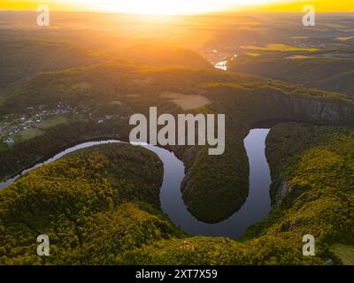 Die Moldau fließt anmutig durch die üppigen grünen Hügel, während die Sonne untergeht, und wirft einen goldenen Glanz über die Landschaft in Tschechien. Diese ruhige Szene fängt das Wesen des Sommers ein. Stockfoto