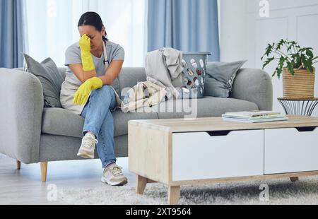 Frustriert, müde und Frau auf der Couch mit Wäsche, unordentlichem Wohnzimmer und Putzkleidung. Hausarbeit, Handschuhe und müde Putzfrau auf dem Sofa in der Wohnung Stockfoto