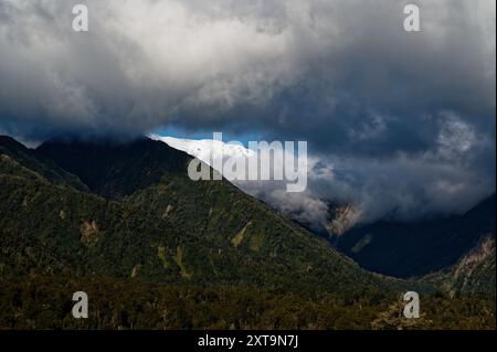 Niedrige Wolken werfen ihren Schatten auf die Hügellandschaft, während schneebedeckte Berge durch eine Lücke ragen. Stockfoto