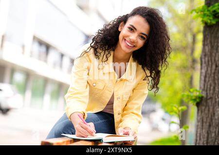 Foto eines jungen positiven Mädchens mit gewellter Haarstyling-Studentin schreibt Notizen im Copybook in gelber trendiger Jacke draußen, die sich auf die Untersuchung vorbereitet Stockfoto