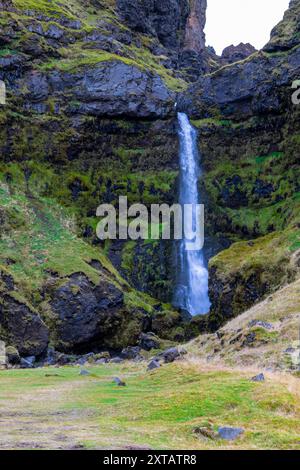 Ein Wasserfall ist im Vordergrund einer felsigen Klippe zu sehen. Der Wasserfall ist umgeben von üppig grünem Gras und Felsen. Die Szene ist ruhig und friedlich Stockfoto