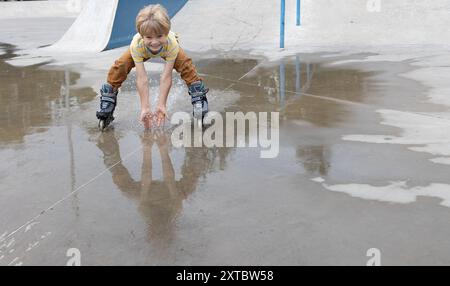 Der Junge reitet nach dem Regen fröhlich mit Geschwindigkeit auf Rollschuhen durch eine Pfütze und spritzt mit seinen Händen. Stimmung zu verwöhnen, gesunder Lebensstil. Inte Stockfoto