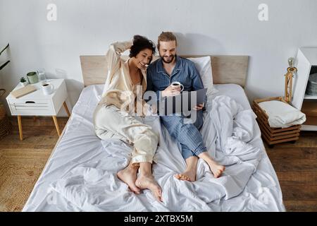 Ein liebevolles Paar kuschelt sich im Bett, genießt Kaffee und benutzt einen Laptop. Stockfoto