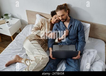 Ein Paar im Pyjama entspannt sich im Bett, während der Mann einen Laptop benutzt. Stockfoto