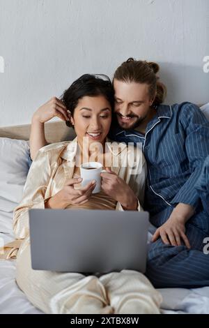 Ein liebevolles Paar entspannt sich im Bett und genießt Kaffee und einen Laptop. Stockfoto