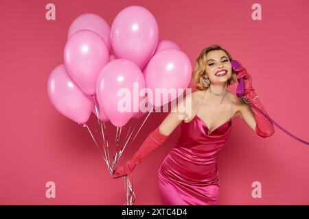 Eine Frau in einem leuchtend rosa Kleid hält einen Haufen pinkfarbene Ballons und redet auf einem pinkfarbenen Telefon. Stockfoto