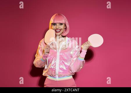 Frau in rosa Jacke und Perücke hält Tischtennispaddles und lächelt vor rosa Hintergrund. Stockfoto