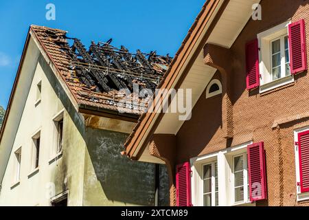 Ein ausgebranntes, verkohltes Dachgebinde klafft neben einem unbeschädigten Nachbargebäude mit roten Fensterläden in der Gotthardstrasse in Andermatt, Uri, Schweiz offen. Stockfoto