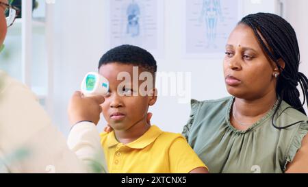 Junge in der Arztpraxis, der die Temperatur überprüft, mit besorgter Mutter an seiner Seite Stockfoto