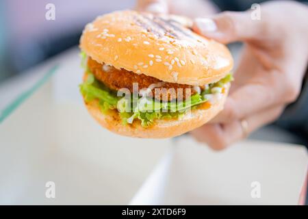 Eine Nahaufnahme einer Hand, die einen Sesambrötchen-Burger hält, gefüllt mit knusprigem gebratenem Huhn, frischem Salat und Mayonnaise, vor einem verschwommenen Hintergrund Stockfoto