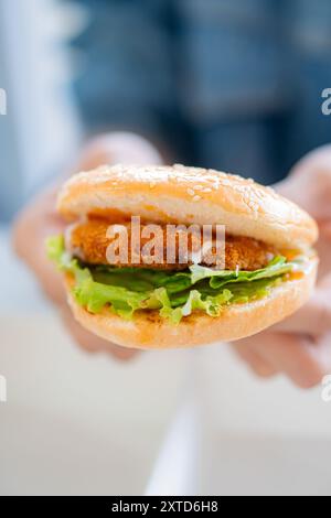 Eine Nahaufnahme einer Hand, die einen Sesambrötchen-Burger hält, gefüllt mit knusprigem gebratenem Huhn, frischem Salat und Mayonnaise, vor einem verschwommenen Hintergrund Stockfoto