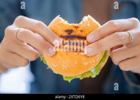 Eine Nahaufnahme einer Hand, die einen Sesambrötchen-Burger hält, gefüllt mit knusprigem gebratenem Huhn, frischem Salat und Mayonnaise, vor einem verschwommenen Hintergrund Stockfoto