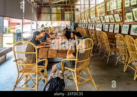 Die Leute sitzen und essen an einem großen Holztisch. Auf der einen Seite werden Aquarelle zum Verkauf im hin Bus Depot in George Town, Penang, ausgestellt Stockfoto