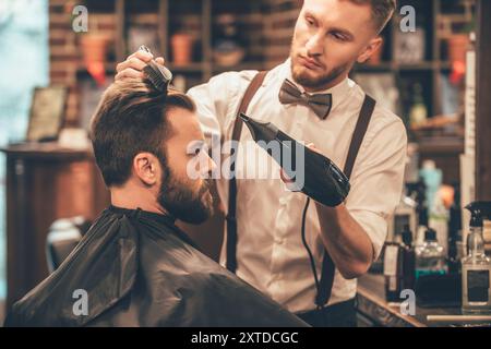 Neue Frisur. Seitenansicht eines jungen bärtigen Mannes, der im Friseur mit Haartrockner gepflegt wurde, während er im Stuhl im Friseur sitzt Stockfoto