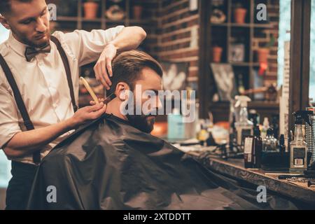 Das Haar sieht magisch aus. Junger bärtiger Mann, der vom Friseur mit Rasiermesser geschnitten wird, während er im Stuhl im Friseur sitzt Stockfoto