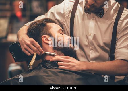 Das Haar sieht magisch aus. Nahaufnahme eines jungen bärtigen Mannes, der vom Friseur im Friseur mit einem Rasierer rasiert wird Stockfoto