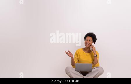 Selbstbewusste Frau, die mit dem Kunden über das Handy spricht, während sie mit Laptop auf weißem Hintergrund sitzt Stockfoto