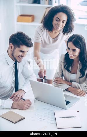 Werfen Sie einen Blick auf diese Tabelle! Blick von oben auf die junge schöne afrikanische Frau, die mit einem Lächeln auf den Laptop zeigt, während sie mit ihren Kollegen im Büro steht Stockfoto