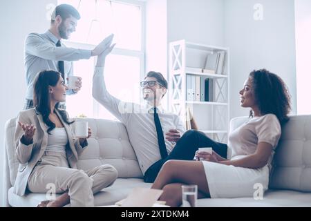 Sie verdienen eine kleine Pause. Zwei gutaussehende Männer geben High-Five und halten Kaffeetassen mit einem Lächeln, während sie mit ihrer Schönheit auf der Couch sitzen Stockfoto