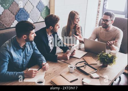 Was sollten wir als Nächstes tun? Blick von oben auf einen jungen, gutaussehenden Mann in Brille, der mit seinen Kollegen etwas mit einem Lächeln bespricht und dabei sitzt Stockfoto