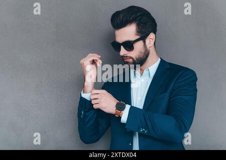 Alles muss perfekt sein. Nahaufnahme eines jungen, gutaussehenden Mannes in einer Sonnenbrille, die den Ärmel an seiner Jacke anpasst, während er vor grauem Hintergrund steht Stockfoto